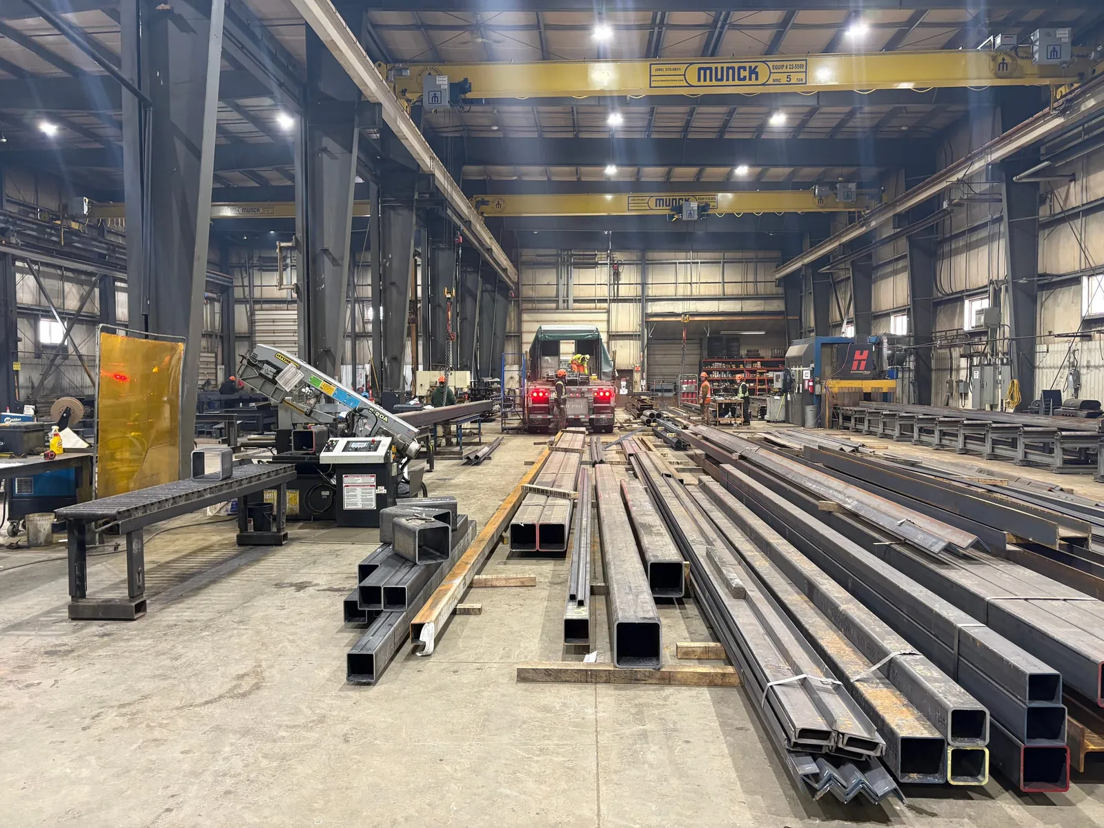 Inside the Portex shop: two yellow overhead bridge cranes span the bay above lengths of square steel tube, a band saw and a flatbed truck being loaded at the far door.