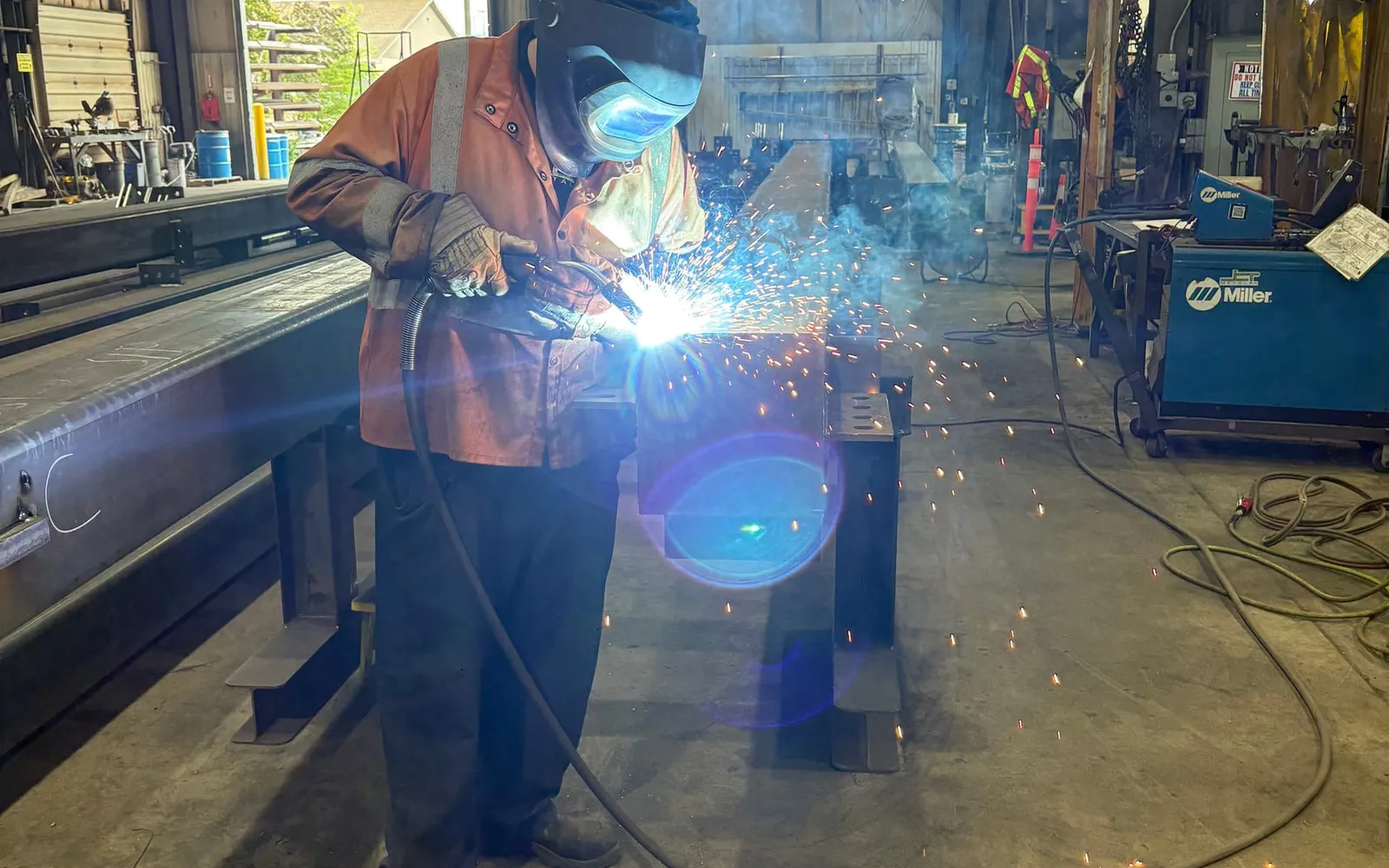 A welder in a helmet and protective jacket welds a steel member on stands, sparks flying, inside the shop.
