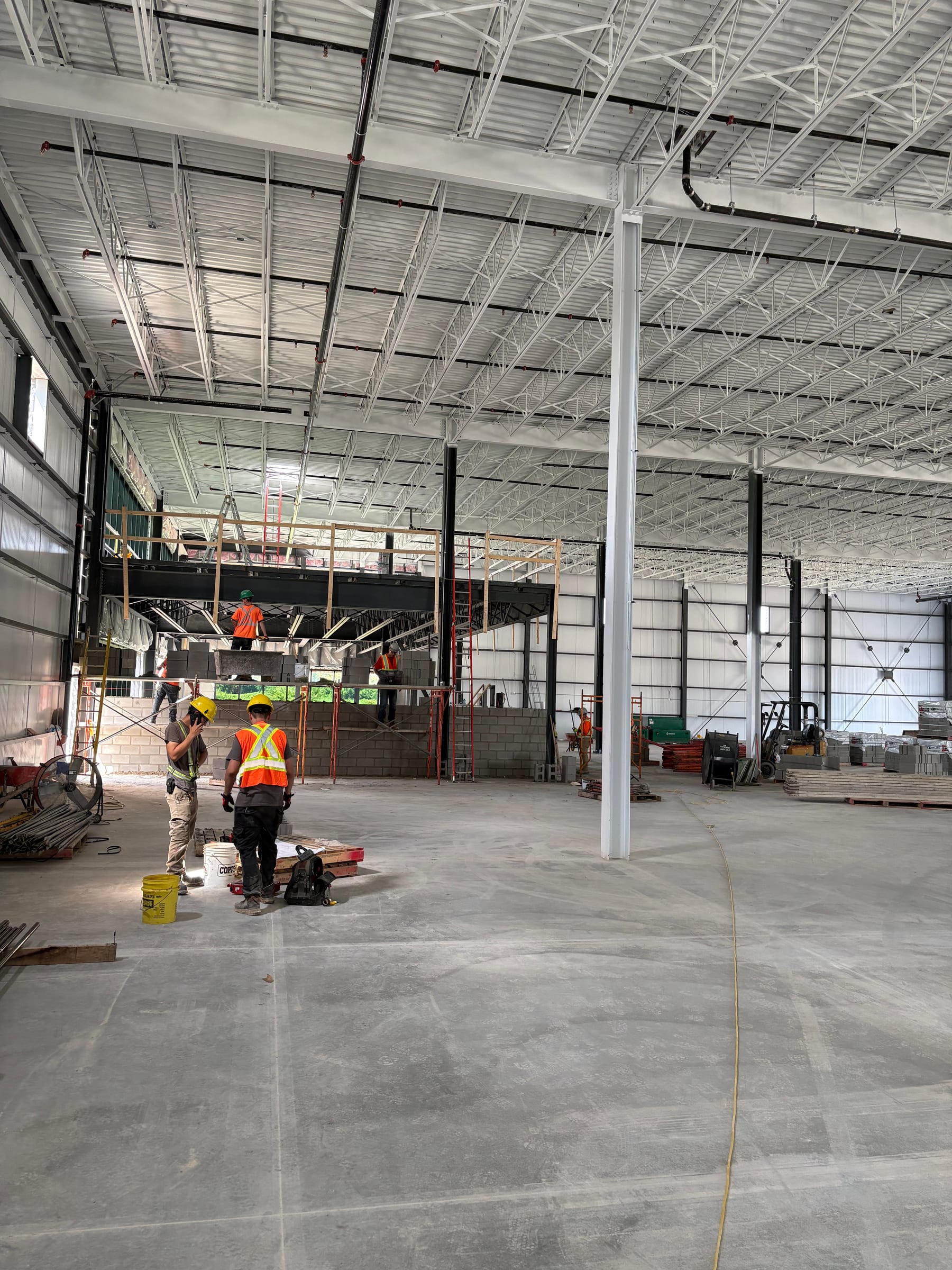 The Phase 1 interior during fit-out: white steel columns and an insulated roof on open-web joists, with masons on scaffolding at the mezzanine at the back.