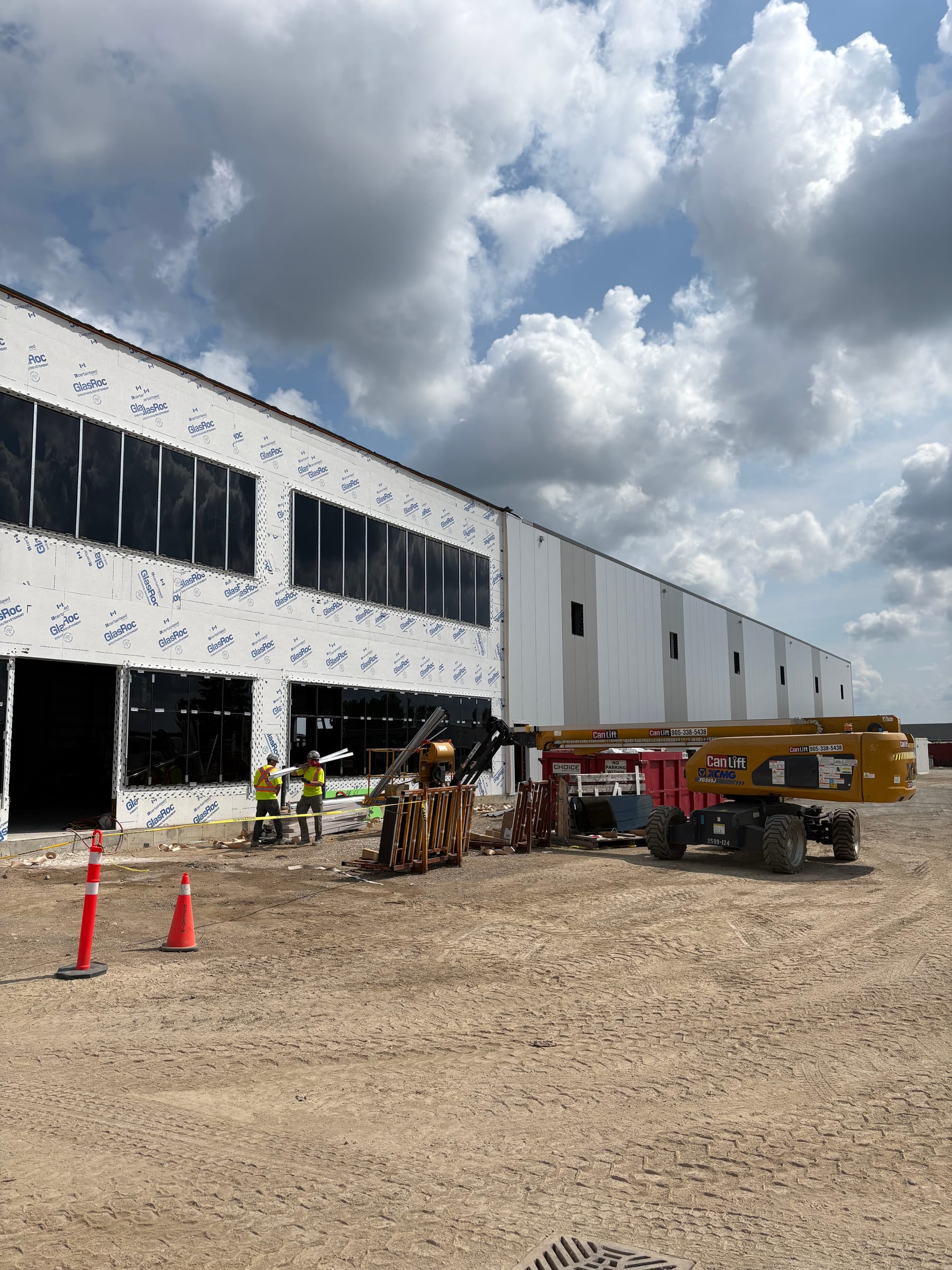The clad front of the Phase 1 building, with rows of window openings in the two-storey office front, a boom lift and safety cones on the gravel in front.