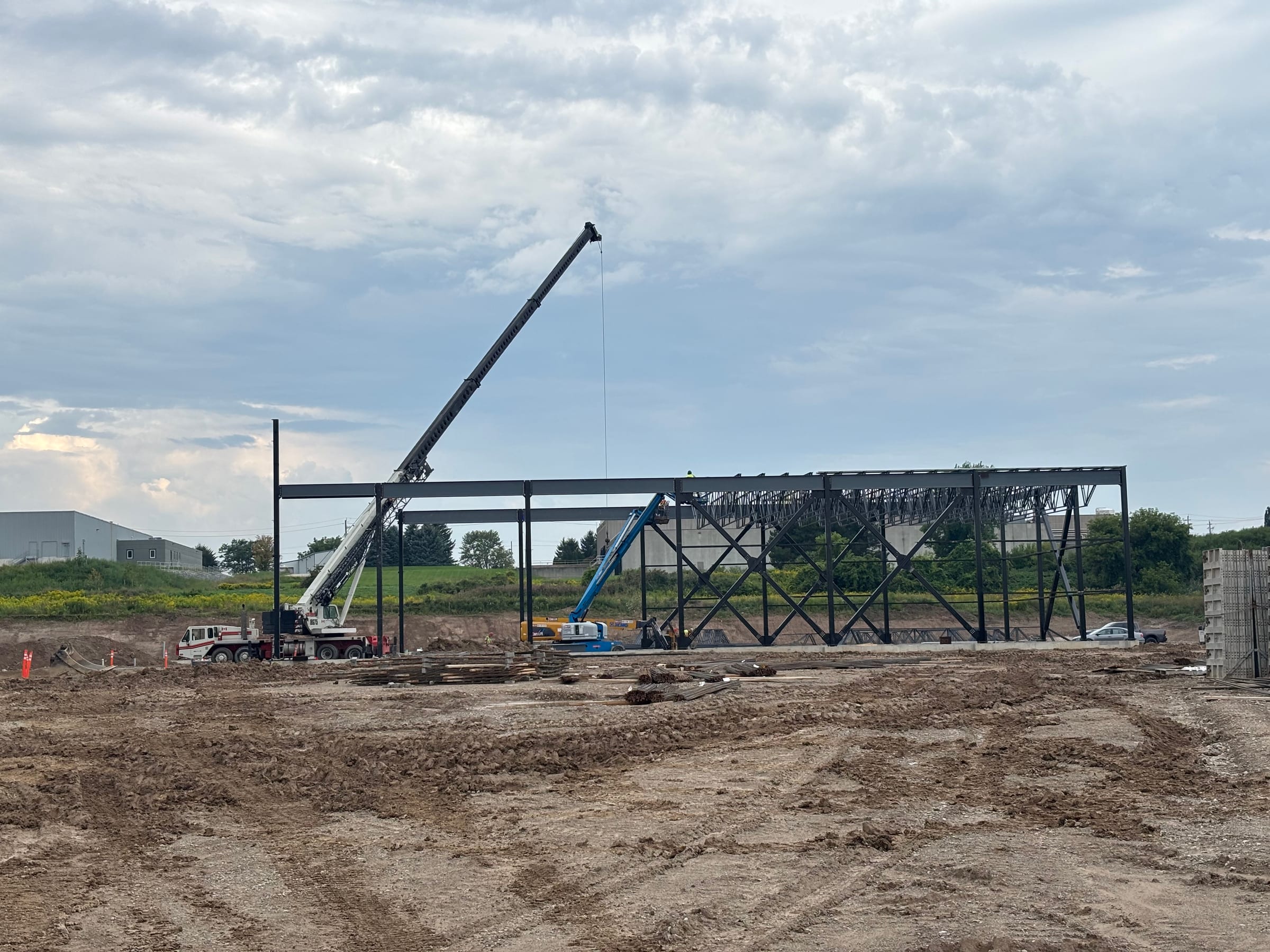 A long braced steel frame across a muddy site under an overcast sky, with a crane lifting steel at the left and a boom lift beside the frame.