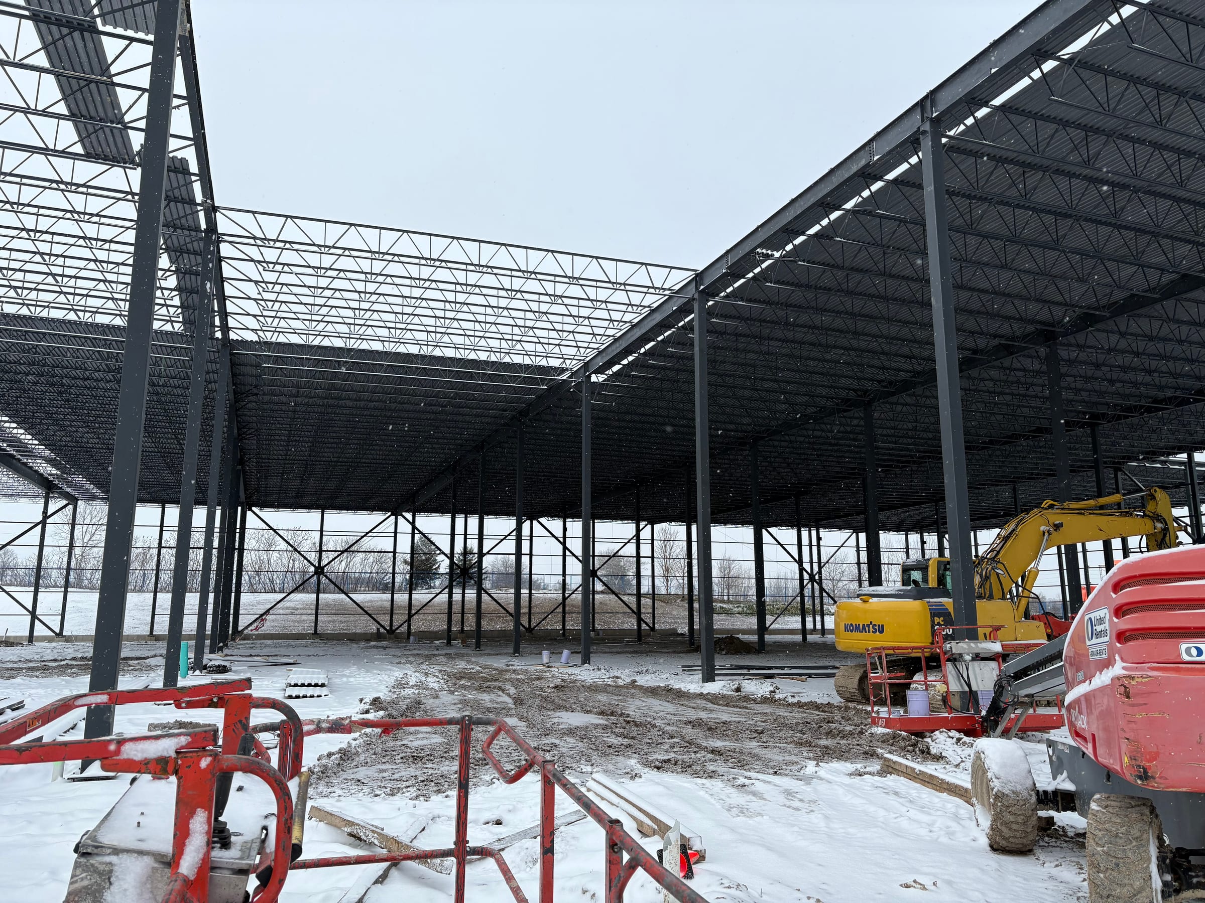 A huge field of open-web roof joists on steel columns going up over snow-covered ground, with an excavator and a boom lift on site.