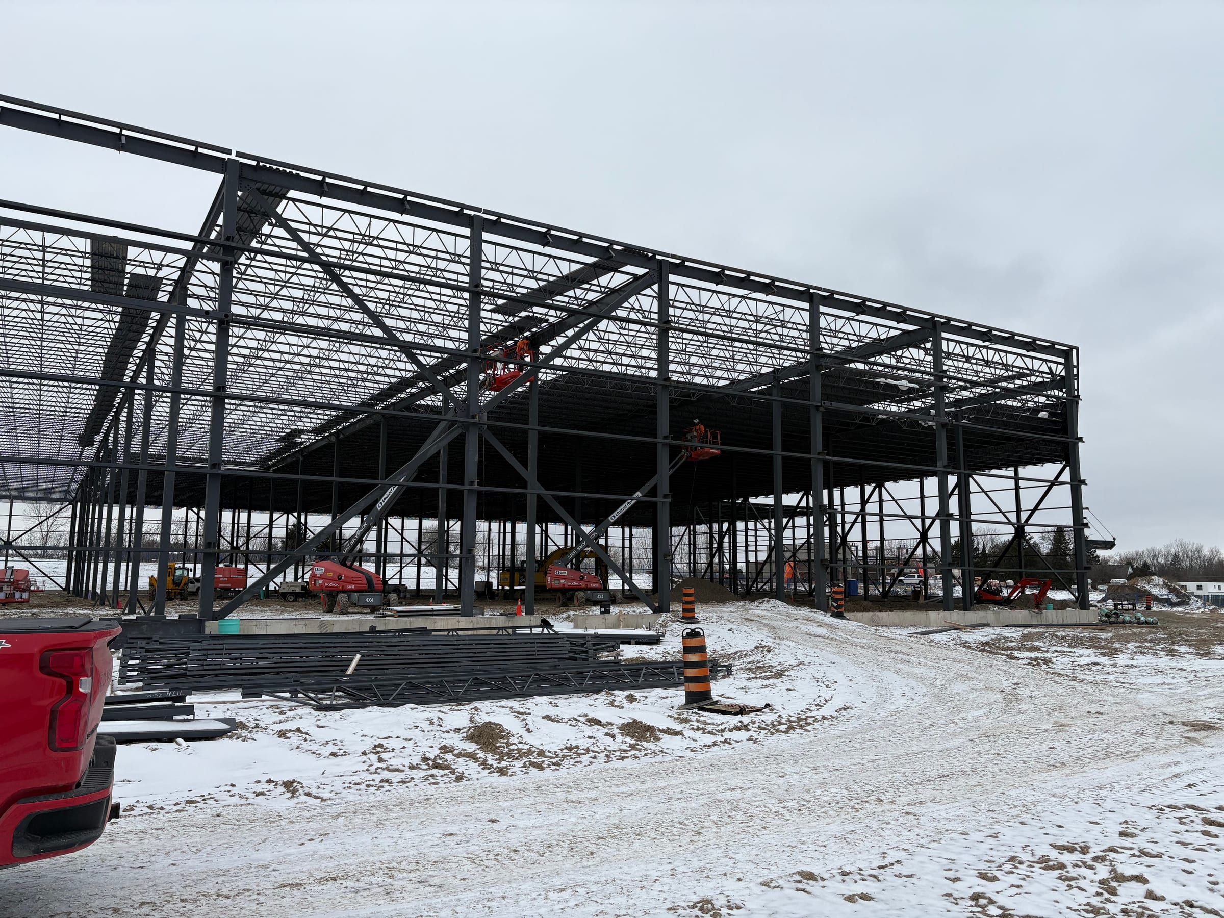 The steel frame and roof joists under erection in snow, with braced bays along the side, boom lifts inside and steel laid out on the ground.
