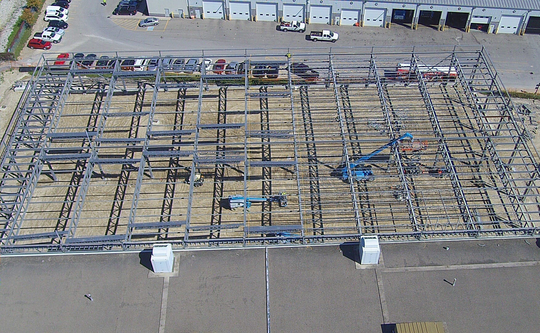 Looking down on the steel frame from above: rows of open-web joists between beams, with boom lifts working and parked buses along the garage.