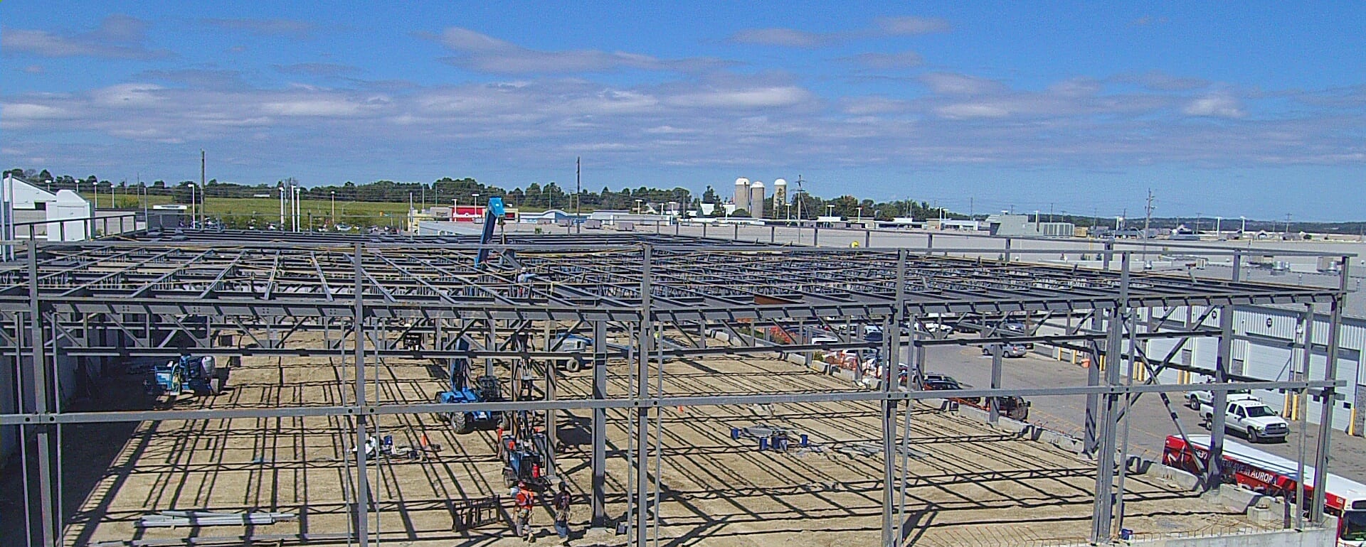 A low aerial view across the steel frame and roof joists on a sunny day, with a telehandler and workers inside the frame and a transit bus parked at the right.